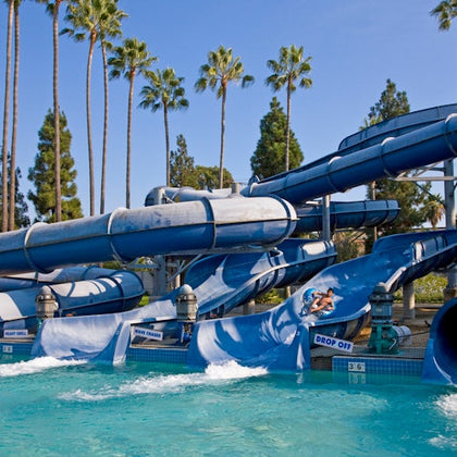 A water park with blue water slides, a pool at the bottom, and palm trees in the background under a clear sky.
