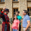 A uniformed guard gestures while speaking to a family of three outside a stone building with arched windows.