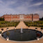 A large, ornate red-brick palace with a symmetrical garden and central fountain under a partly cloudy sky.