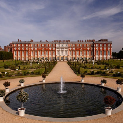 A large, ornate red-brick palace with a symmetrical garden and central fountain under a partly cloudy sky.