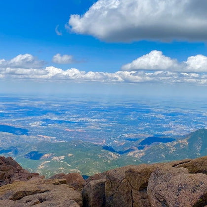 A view from a rocky mountain summit overlooking a vast landscape with hills, a city, and a sky filled with clouds.
