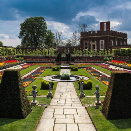 A formal garden with a central fountain, symmetrical flowerbeds, hedges, statues, and a brick building in the background under a cloudy sky.