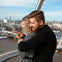 A couple embraces inside a ferris wheel pod, overlooking a river and a cityscape with bridges and buildings in the background.