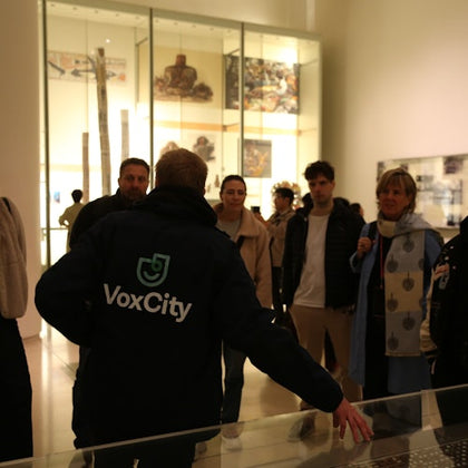 A group of people in a museum setting, with a guide wearing a 'VoxCity' jacket explaining exhibits to the group.