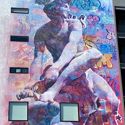 A colorful mural on a building featuring a large statue-like figure overlaid with vibrant graffiti art and patterns.
