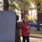 A woman in a red jacket stands smiling with arms crossed next to a statue in a park during autumn with trees and cars nearby.