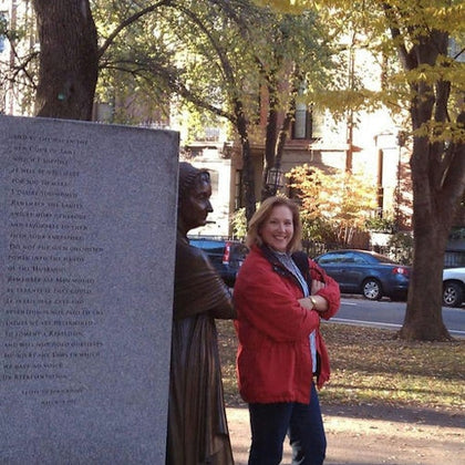 A woman in a red jacket stands smiling with arms crossed next to a statue in a park during autumn with trees and cars nearby.