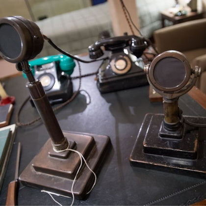 Two vintage microphones and telephones are on a desk in a retro office setting.