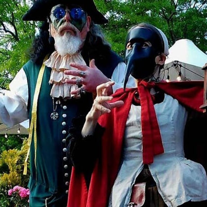 Two people in elaborate costumes, one in a green outfit with a hat and sunglasses, the other in a mask and red cape, outdoors.
