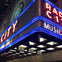 Neon signs of Radio City Music Hall's marquee, featuring blue and red lights with labeled lanes below.