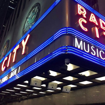 Neon signs of Radio City Music Hall's marquee, featuring blue and red lights with labeled lanes below.
