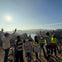 Five people in helmets stand with bicycles, raising hands, overlooking a scenic landscape with hills and a lake under a sunny sky.