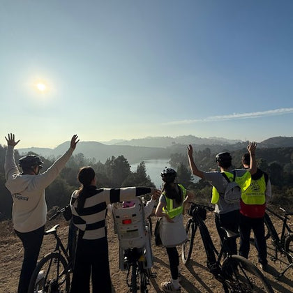 Five people in helmets stand with bicycles, raising hands, overlooking a scenic landscape with hills and a lake under a sunny sky.