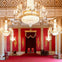 An ornate room with red walls, gold details, chandeliers, and two throne-like chairs under a canopy.