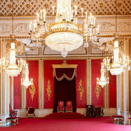 An ornate room with red walls, gold details, chandeliers, and two throne-like chairs under a canopy.