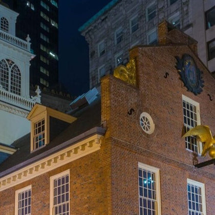A historic red brick building with golden ornaments and a clock, surrounded by modern and classical architecture at night.