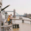 Two people stand on a ship's deck overlooking the Thames River with Tower Bridge visible in the background.