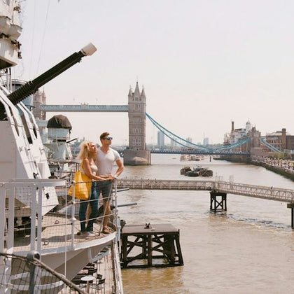 Two people stand on a ship's deck overlooking the Thames River with Tower Bridge visible in the background.