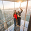 Three people standing on a glass platform overlooking a city skyline, taking a selfie with the sun setting in the background.