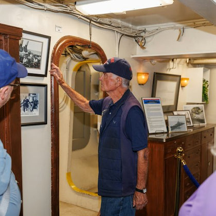 A man wearing a cap points to framed photos on a wall inside a ship's interior, with a porthole and various displays visible.