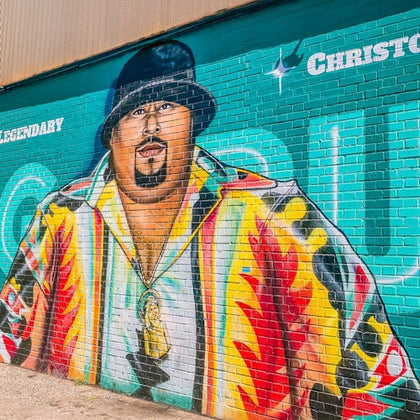 Colorful mural of a man wearing a vibrant shirt and black hat, with text saying "Legendary" and "Christopher" on a brick wall.