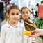 Two children watch a large brown butterfly with eye-like patterns on its wings, perched on an orange slice held by a hand.