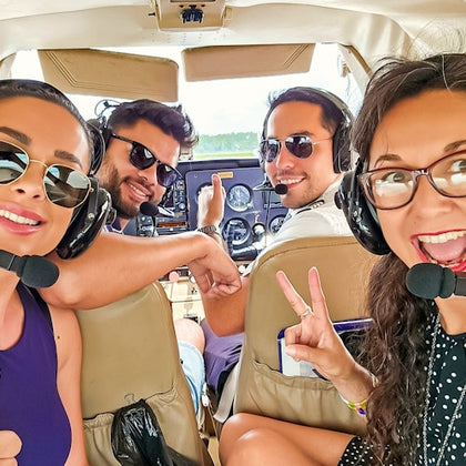 Four smiling people wearing headsets inside a small airplane cockpit, two seated in front and two in the rear, making peace signs.