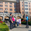 A group of people stand outside a historic brick building with flowers and greenery in the foreground.