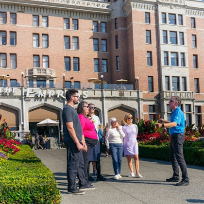 A group of people stand outside a historic brick building with flowers and greenery in the foreground.