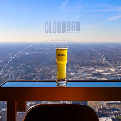 A tall glass of yellow drink on a table with a cityscape view through a large window, labeled 'CloudBar at 360 Chicago.'