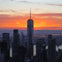 A city skyline at sunset with prominent skyscrapers, including a tall building with a spire, silhouetted against an orange sky.
