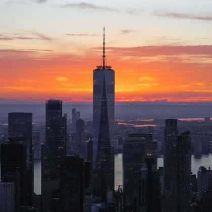 A city skyline at sunset with prominent skyscrapers, including a tall building with a spire, silhouetted against an orange sky.