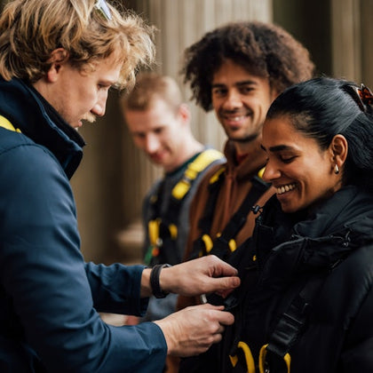 A group of people preparing for an activity, with one person assisting another with safety harnesses, all smiling.