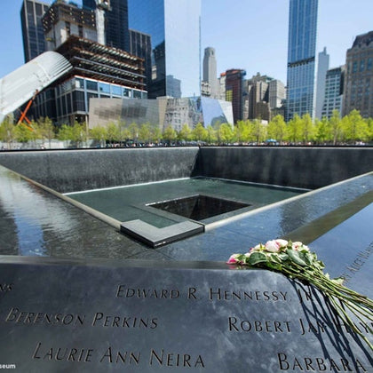 Memorial water fountain with names inscribed, surrounded by skyscrapers. Pink and white flowers are placed on the edge.