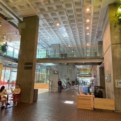 Spacious atrium with high ceiling, featuring modern decor, large windows, and people sitting at tables and walking.