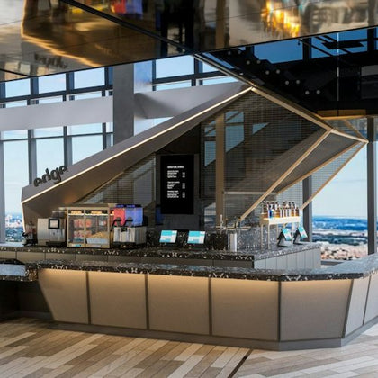 A modern cafe counter with beverage dispensers, menu screens, and a geometric backdrop, located in a high-rise setting.