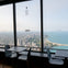 Tables and chairs by large windows overlooking a cityscape and a coastline with buildings extending along a sandy beach.
