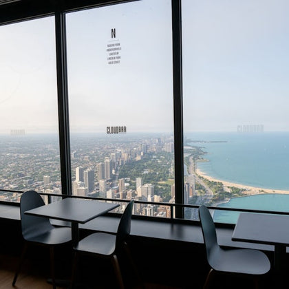 Tables and chairs by large windows overlooking a cityscape and a coastline with buildings extending along a sandy beach.