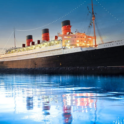 An illuminated ocean liner with three red and black smokestacks is docked at twilight, reflected in calm water.