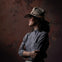 A person wearing a wide-brimmed hat and a buttoned shirt, standing against a textured background, looking to the side.