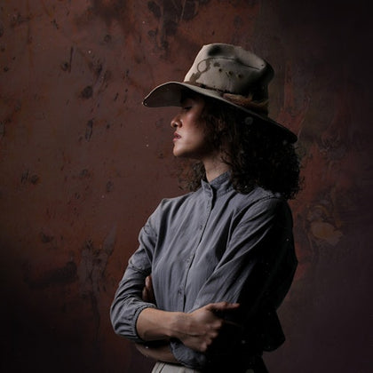 A person wearing a wide-brimmed hat and a buttoned shirt, standing against a textured background, looking to the side.