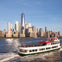 A sightseeing cruise boat on the water with a group of people on board, with the New York City skyline in the background.
