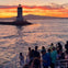 A group of people on a boat watch a sunset over a lighthouse on a rocky outcrop with hills in the background.