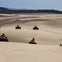People riding red ATVs across vast sandy dunes under a clear sky. Forested hills are visible in the distance.