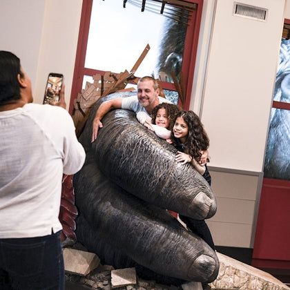 A person takes a photo of three people posing with a large sculpture of a gorilla's hand in an indoor space.
