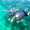 An adult and child snorkeling in clear, blue-green water. The child has a purple pool noodle for support.
