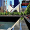 Modern cityscape with a futuristic building, a reflecting pool, trees, and people along a walkway under a clear blue sky.