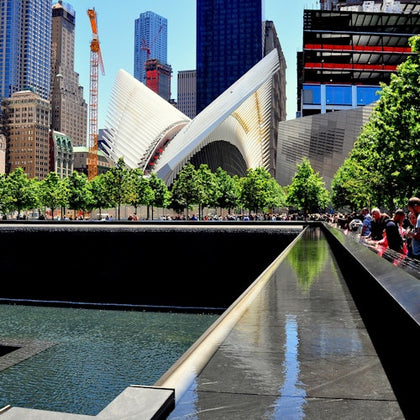Modern cityscape with a futuristic building, a reflecting pool, trees, and people along a walkway under a clear blue sky.