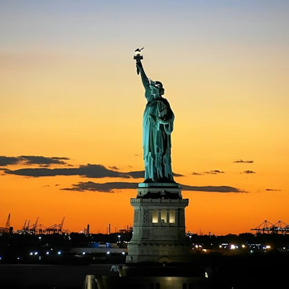 The Statue of Liberty against a vibrant orange sunset sky, with a silhouetted cityscape and cranes in the background.