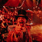 A circus ringmaster in an ornate outfit enthusiastically gestures towards the camera, with a crowd and lights in the background.